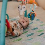 Cute baby lying on a play mat wearing a onesie and interacting with hanging toys.